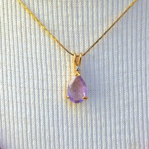 May include: A gold-toned necklace with a teardrop-shaped, light purple gemstone pendant. The pendant is suspended from a delicate chain. The background is a white fabric and a red sweater.
