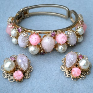 May include: A vintage jewelry set featuring a gold-tone hinged bracelet and matching earrings. The set is adorned with pink, white, and lavender-colored stones, faux pearls, and small purple accents. The earrings have a filigree design.