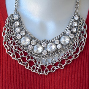 May include: A silver-toned statement necklace featuring multiple layered metal chains and a series of round, clear faceted crystals set in circular frames. The design incorporates a mix of large and small stones arranged in a wide, collar-style pattern.