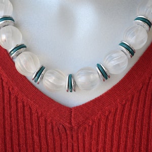 May include: A necklace featuring large, spherical, frosted white beads with a ribbed texture, alternating with smaller, flat, teal-colored circular spacers and thin silver-toned metal discs, resting against a ribbed, V-neck red garment.