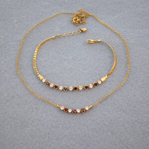 May include: A gold-toned jewelry set featuring a necklace and bracelet. The necklace and bracelet have a series of alternating red and clear gemstones. The necklace has a delicate chain, and the bracelet has a textured band.