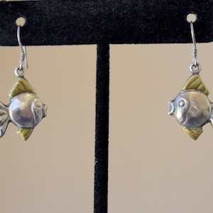 May include: Silver fish-shaped earrings with gold-colored fins and accents. The earrings are suspended from silver hooks. The fish have a rounded body and detailed fins, creating a whimsical, aquatic-themed jewelry piece.