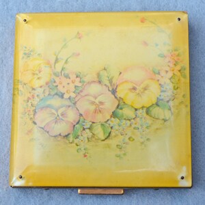 May include: A square, yellow compact with a floral design. The compact features a painted image of pansies in shades of yellow, pink, and purple, surrounded by green leaves and small flowers. The compact has a gold-colored metal clasp and corner accents.