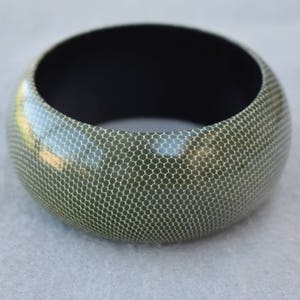 May include: A wide, round bangle bracelet with a textured surface. The bracelet has a dark interior and a light green exterior with a repeating honeycomb pattern. The jewelry is made of a glossy material.