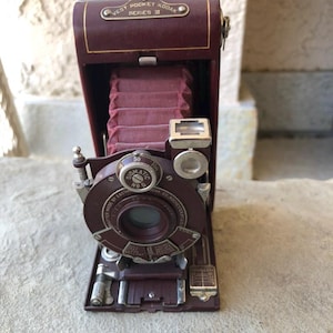 May include: A vintage maroon Vest Pocket Kodak Series III camera. The camera has a collapsible bellows and a circular lens with dials. The top of the camera has the text "Vest Pocket Kodak Series III" in gold lettering.