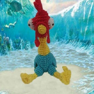 May include: A crocheted rooster toy with red, yellow, and blue yarn. The rooster is sitting on a white sandy beach with blue water in the background. The word "MOANA" is written in the top of the image.