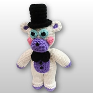 May include: A handmade crochet bear with a black top hat, bow tie, and purple accents. The bear has white fur, blue eyes, pink cheeks, and a purple muzzle. The bear's feet and ears are also trimmed in purple.