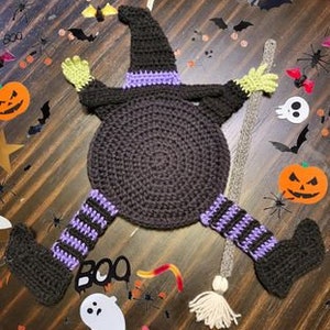 May include: A crocheted witch doll with a brown hat, purple and white striped legs, and a brown broom. The doll is lying on a table with a brown surface. The doll is surrounded by Halloween decorations, including pumpkins, bats, and stars.