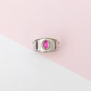 May include: A silver ring featuring a pink oval gemstone set in the centre. The ring has a wide band with a textured surface.