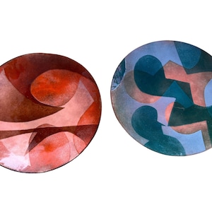 May include: Two round ceramic plates with abstract geometric designs. One plate is red, brown, and white. The other plate is blue, green, and pink.