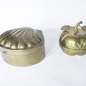 May include: Two brass trinket boxes. One is shaped like a scallop shell, the other like an apple with a stem and leaves. Both boxes have a shiny, metallic finish and are likely vintage decorative items.