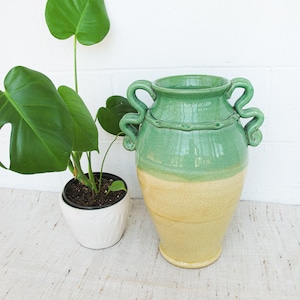 May include: A green and yellow ceramic vase with two handles. The vase is sitting on a white surface with a green plant in a white pot.