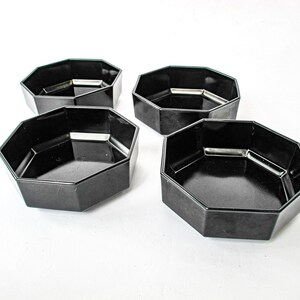 May include: Four black, octagonal serving bowls are arranged on a white surface. The bowls have a glossy finish and a simple, modern design. They are likely made of plastic or a similar material.