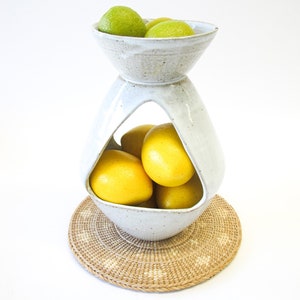 May include: A white ceramic fruit bowl with a unique hourglass shape, holding green limes in the top bowl and yellow lemons in the bottom. The bowl sits on a round, woven placemat, creating a stylish centerpiece.