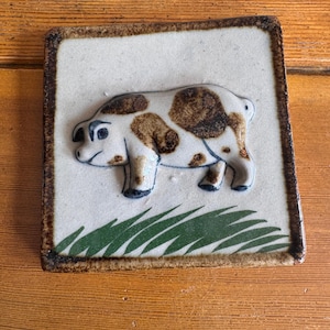 May include: A ceramic square tile featuring a white pig with brown spots. The pig is raised from the surface and stands on a base of green grass. The tile has a brown border and is a decorative item.