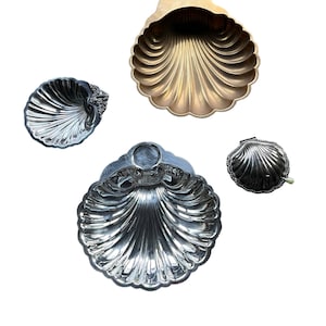 May include: Four decorative seashell-shaped dishes in various sizes and colors. The shells are made of metal and have a ribbed texture. One is gold, two are silver, and one is a smaller silver shell with a decorative edge.