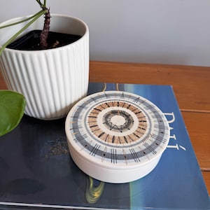 May include: A round, white ceramic trinket box with a patterned lid. The lid features a concentric design in shades of brown, black, and blue. A white potted plant with green leaves and a brown stem is in the background.