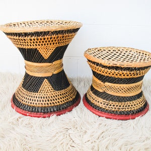 May include: Two woven rattan stools. The larger stool has a hourglass shape with black and natural woven patterns, a red base, and a woven top. The smaller stool has a similar design. The stools are on a white background.