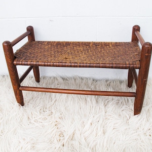 Könnte beinhalten: Eine Holzbank mit einem geflochtenen Sitz. Die Bank hat ein schlichtes Design mit vier Beinen und einer einzigen Strebe. Der Sitz ist aus einem natürlichen geflochtenen Material, möglicherweise Weide oder Rattan.