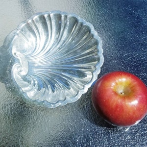 May include: A silver, shell-shaped dish with a decorative design sits on a blue and white textured surface next to a red apple.