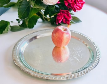 May include: A red apple sits on a silver serving tray with a decorative edge. The tray is on a white table with a bouquet of pink and red roses in the background.