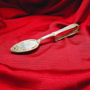 Puede incluir: Pinzas de azúcar de plata con un diseño adornado, sobre una superficie de tela roja.