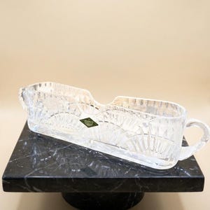 May include: A clear, elongated glass serving dish with a scalloped rim and a handle on one side. The glass features a pressed fan-like pattern throughout its body. A small, diamond-shaped green sticker with gold text is attached to the front surface.