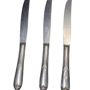 Puede incluir: Tres cuchillos plateados con hojas curvas y mangos ornamentados. Los mangos tienen un diseño decorativo cerca de la base y un detalle en forma de corazón en la parte inferior. Los cuchillos están dispuestos verticalmente sobre un fondo blanco.