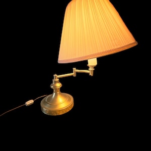 May include: A brass-toned adjustable desk lamp with a pleated, peach-colored lampshade. The lamp has a round base and an articulated arm. The lampshade is trimmed with a decorative border. The lamp is plugged into a wall outlet.