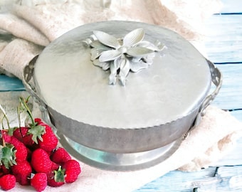 May include: A silver-toned metal serving dish with a lid, featuring a floral embellishment on top. The dish has handles and sits on a small base. Red raspberries and greenery are arranged nearby on a light-colored cloth, set against a blue wooden surface.