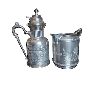 May include: A silver-colored antique coffee pot and creamer set. The coffee pot has a tall, ornate design with a lid and handle. The creamer has a floral design and a handle. Both pieces are made of metal.