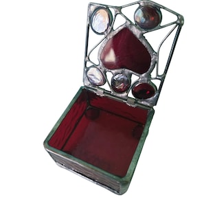 Puede incluir: Caja de vidrio manchado abierta con tapa con bisagras. La tapa presenta una pieza de vidrio roja en forma de corazón rodeada de detalles de vidrio transparente e iridiscente. El interior de la caja está forrado con vidrio rojo.