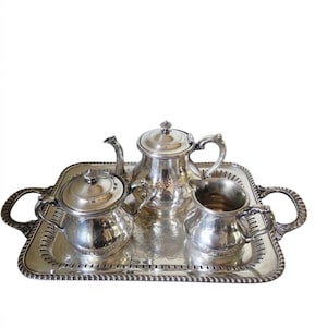 May include: A silver-plated tea service set consisting of a teapot, a lidded sugar bowl, and a creamer, all arranged on a rectangular silver-plated serving tray with ornate handles and a decorative pierced border with a rope-style edge.