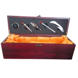 May include: A rectangular dark wood wine accessory box with a hinged lid, featuring a dark red finish and a gold-coloured latch. The interior is lined with bright orange fabric and contains a black foam insert holding a metal ring, stopper, pourer, thermometer, and a corkscrew.