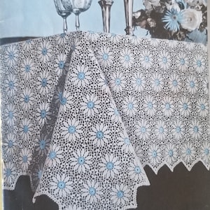 May include: A white crocheted tablecloth with a scalloped edge and a repeating pattern of blue flowers. The tablecloth is laid out on a dark surface with glasses and candlesticks in the background.