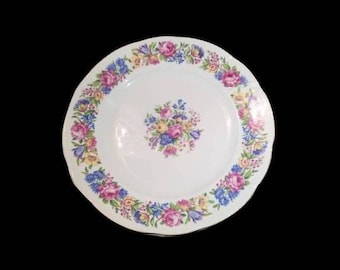 Puede incluir: Un plato de cerámica blanco con un diseño floral. El plato tiene un borde festoneado y presenta un borde de flores rosas, azules y amarillas. El centro del plato tiene un grupo de flores en los mismos colores.