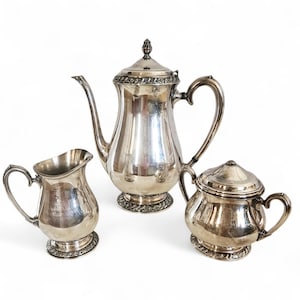 May include: A silver tea set including a coffee pot with a lid, a creamer, and a sugar bowl with a lid. The set has ornate detailing around the base and the lids. The pieces are reflective and sit on a white background.