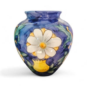 May include: A hand-painted ceramic vase with a blue background and a large white daisy with a yellow center. Green leaves and yellow accents surround the flower. The vase is a decorative home decor item.