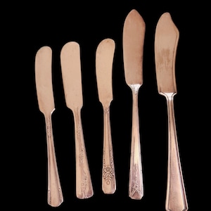 Puede incluir: Cinco cuchillos de mantequilla plateados antiguos con diferentes diseños de mango. Las hojas son anchas y redondeadas, y los mangos presentan diseños florales y geométricos. Los cuchillos están colocados sobre un fondo negro.