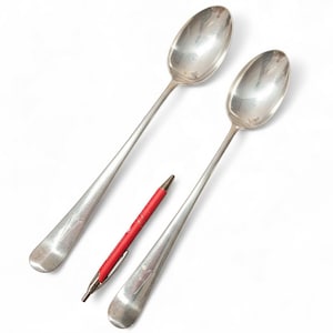 May include: Two silver spoons with long handles and oval bowls are displayed on a white background. A red pen with a silver clip rests between the spoons. The spoons have a decorative design near the bowl.