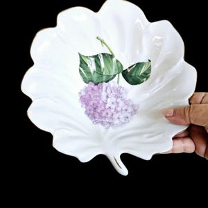 May include: White porcelain leaf-shaped dish with a gold rim. The dish features a painted design of a purple hydrangea flower with green leaves.