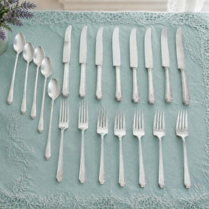 May include: A set of silver-toned flatware consisting of eight dinner knives, seven dinner forks, and five spoons arranged on a light blue tablecloth. The cutlery features a simple, slender handle design with a subtle decorative detail at the base.