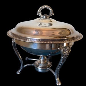 May include: A silver-toned chafing dish with a polished domed lid featuring a decorative handle. The round vessel rests on a tripod stand with ornate, scrolled legs and includes a central burner component underneath for heating food contents.