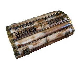 Puede incluir: Una caja rectangular de estilo antiguo, con forma cilíndrica. Está hecha de materiales de color marrón claro y crema, con intrincados calados florales y detalles en latón. La caja tiene una tapa con bisagras y un cierre de broche.