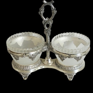 May include: A double condiment server featuring two frosted glass bowls with scalloped rims, held in a silver-toned metal frame with ornate leaf and vine detailing, a central vertical handle, and decorative feet on a black background.