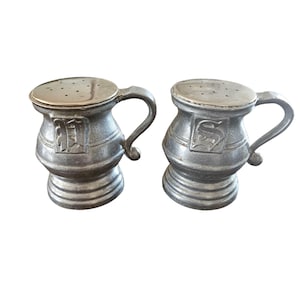 May include: A pair of antique silver-colored salt and pepper shakers. Each shaker has a handle and a lid with small holes. The shakers have a decorative design on the front and a ribbed base. The shakers are likely made of metal.