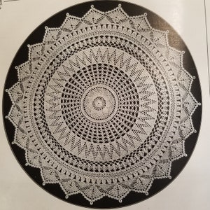 Op de afbeelding: Een wit gehaakt tafellaken met een rond ontwerp en ingewikkelde details. Het tafellaken wordt getoond op een zwarte achtergrond.