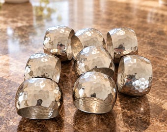 Könnte beinhalten: Ein Set aus sieben silberfarbenen Metall-Serviettenringen, die jeweils eine markante, gehämmert wirkende Oberfläche mit kleinen Vertiefungen aufweisen. Die Ringe sind in einer Gruppe angeordnet und zeigen ein glänzendes Finish.