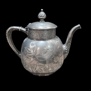 May include: A silver teapot with intricate floral and vine engravings. The teapot features a curved spout, a handle, and a lid with a decorative finial. The design includes detailed leaves, flowers, and grape clusters, set against a black background.