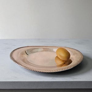 May include: A silver-colored oval serving platter with a rope-like border. A golden-brown fruit sits on the platter, reflecting its shape. The platter is on a light gray surface, creating a simple, elegant presentation.